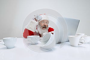 Cropped portrait of senior man wearing Santa Claus costume sitting at the desk full of empty coffee cups and looking on