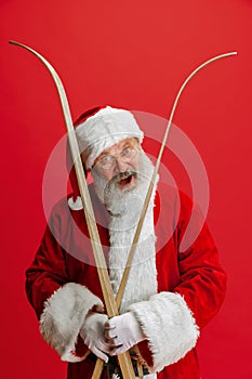 Cropped portrait of joyful senior man wearing Santa Claus costume, holding skis isolated over red background. Winter