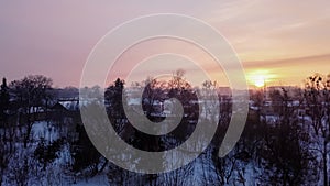 Timelapse, winter sunset over the snow-covered city, dark outlines of bare trees, the roofs of houses, the orange sun