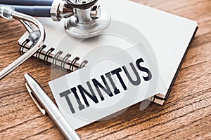 TINNITUS - text on card on wooden table with stethoscope and notepad for medical records