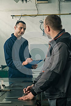 A tinsmith is sketching in his workshop with a colleague engineer who is taking notes