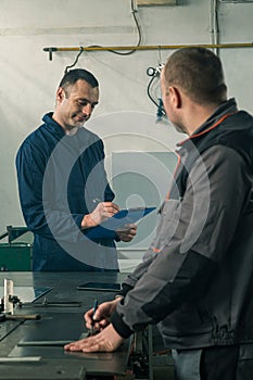 A tinsmith is sketching in his workshop with a colleague engineer who is taking notes