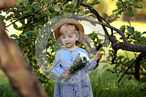 Tiny girl in blue jeans dress, with straw hat, holding bouquet of chamomile, looking to camera, staying near to branch on sunny