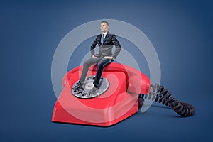 A tiny serious businessman sits patiently on a giant red retro phone with a dial.