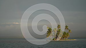 Tiny tropical, uninhabited island in the sea with palm trees and rainbow on the cloudy sky. Vacation and travel concept