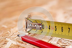 A tip of yellow metal tape measure and red carpenter pencil lying on chip board. Blurred background