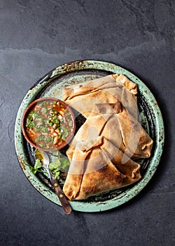 Tipical baked empanadas de pino with pebre sauce on vintage try, black stone background. Traditional chilean food for photo