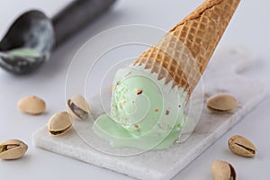 A tipped over pistachio ice cream cone on a small marble slab surrounded by pistachios and a metal scooper in behind.