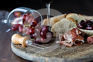 Tipped over wine glass with red grapes spilling out onto a rustic board surrounded by bread, olives and prosciutto.