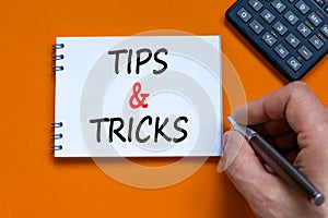 Tips and tricks symbol. Businessman writing words `Tips and tricks` on white note. Black calculator. Beautiful orange background