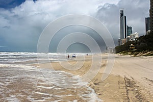 Tire tracks and unidentifiable tourists on beach wading out into surf on stormy day with dark clouds at Gold Coast - Surfers