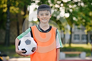 Tired 13-aged soccer player in orange vest holds ball in hand after training on outdoors sport ground at sunny morning.