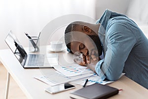 Tired african american freelancer guy sleeping at desk with laptop and papers