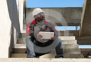 Tired african american worker sits at construction site and holds tablet PC