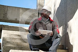 Tired african american worker sits at construction site and holds tablet PC