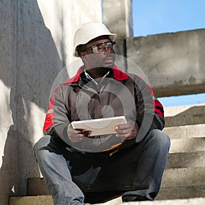 Tired african american worker sits at construction site and holds tablet PC