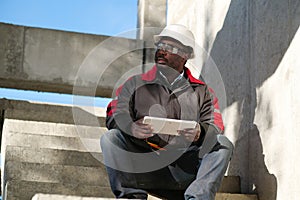 Tired african american worker sits at construction site and holds tablet PC