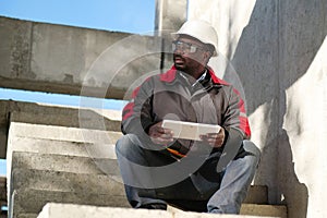 Tired african american worker sits at construction site and holds tablet PC