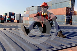 Tired African young worker man wearing safety red vest and helmet, sitting on the floor at logistic container shipping cargo yard
