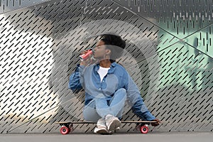 Tired afro american skateboarder girl refreshing after longboarding sit on skate drink soda beverage