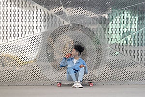 Tired afro american skateboarder girl refreshing after longboarding sit on skate drink soda beverage