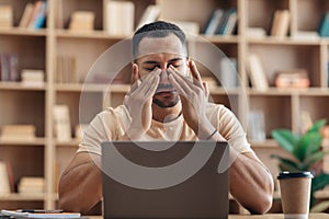 Tired arab freelancer guy sitting at workdesk in front of laptop at home and rubbing his eyes, suffering from burnout