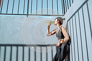Tired athlete leaning against a concrete wall drinks water from a bottle. Young, fit and sporty brunette girl in