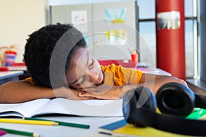 Tired biracial schoolboy sitting at desk and lying on books in classroom