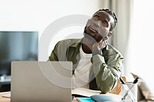 Tired black man suffering from neck spasm while working on laptop and rubbing his muscles, sitting at desk