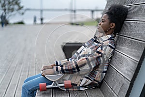 Tired afro skateboarder girl sitting alone on bench in urban open space depressed ponder outdoors