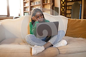 Tired black woman sitting on couch with laptop, staring at screen, feeling exhausted, having dull online job at home