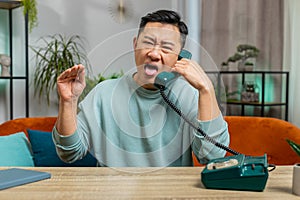 Tired bored Asian man talking on wired telephone exhausted of talk, making silly faces, fooling