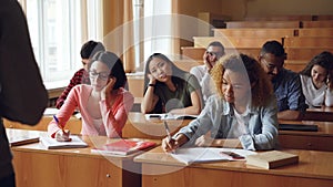 Tired and bored students are listening to teacher and making notes in notebooks sitting at desks in university