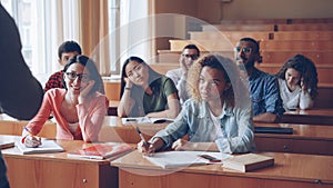 Tired and bored students are listening to teacher and making notes in notebooks sitting at desks in university