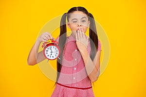 Tired and bored teenager girl with clock alrm, time and deadline. Studio shot isolated on yellow background.