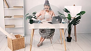 Tired bored young modern woman sits at her desk in light, spacious office. She works with laptop and a notebook. Woman