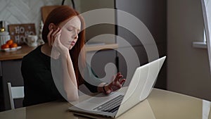 Tired bored young woman typing on laptop keyboard holding hand on chin sitting at table in kitchen room with modern