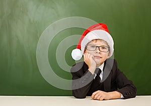Tired boy in red christmas hat sitting near empty green blackboard