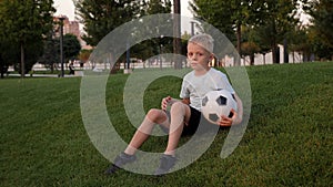 A tired boy is sitting on the green grass in a city park with a soccer ball.