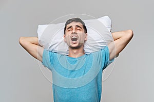 Tired brunet man in a blue tee resting on a white pillow  over grey background.