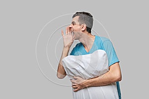 Tired brunet man in a blue tee shout holding white pillow isolated over grey background.