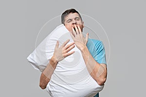 Tired brunet man in a blue tee yawning hugging white pillow isolated over grey background.