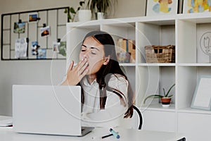 Tired brunette girl yawns tiredly with eyes closed while working with laptop photo