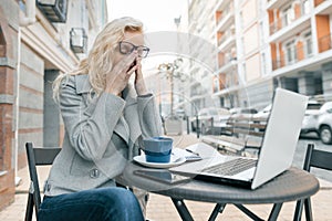 Tired business woman with laptop in outdoor cafe, woman touching her eyes with her glasses off, city street background