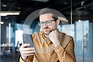 Tired businessman, employee sitting at a desk in an office center and using the phone. Bored and grimacing, he looks at