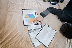 Tired businessman lying on the bed with a pile of graph papers on his hand that was in front of him