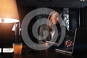 Tired businessman working by the contemporary laptop at night office. Hipster with long hair hold notepad in hand and uses mobile