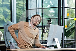 Tired businessman at workplace has severe back and lower back pain, young man working sitting at table with laptop and