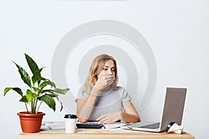 Tired businesswoman yawning while sitting in front of opened laptop, calculating figured and making business report, having sleepl