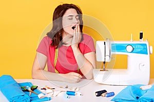 Tired busy seamstress sits at table with sewing machine, closes her eys, yawns, having many things to do, wearing casual red t